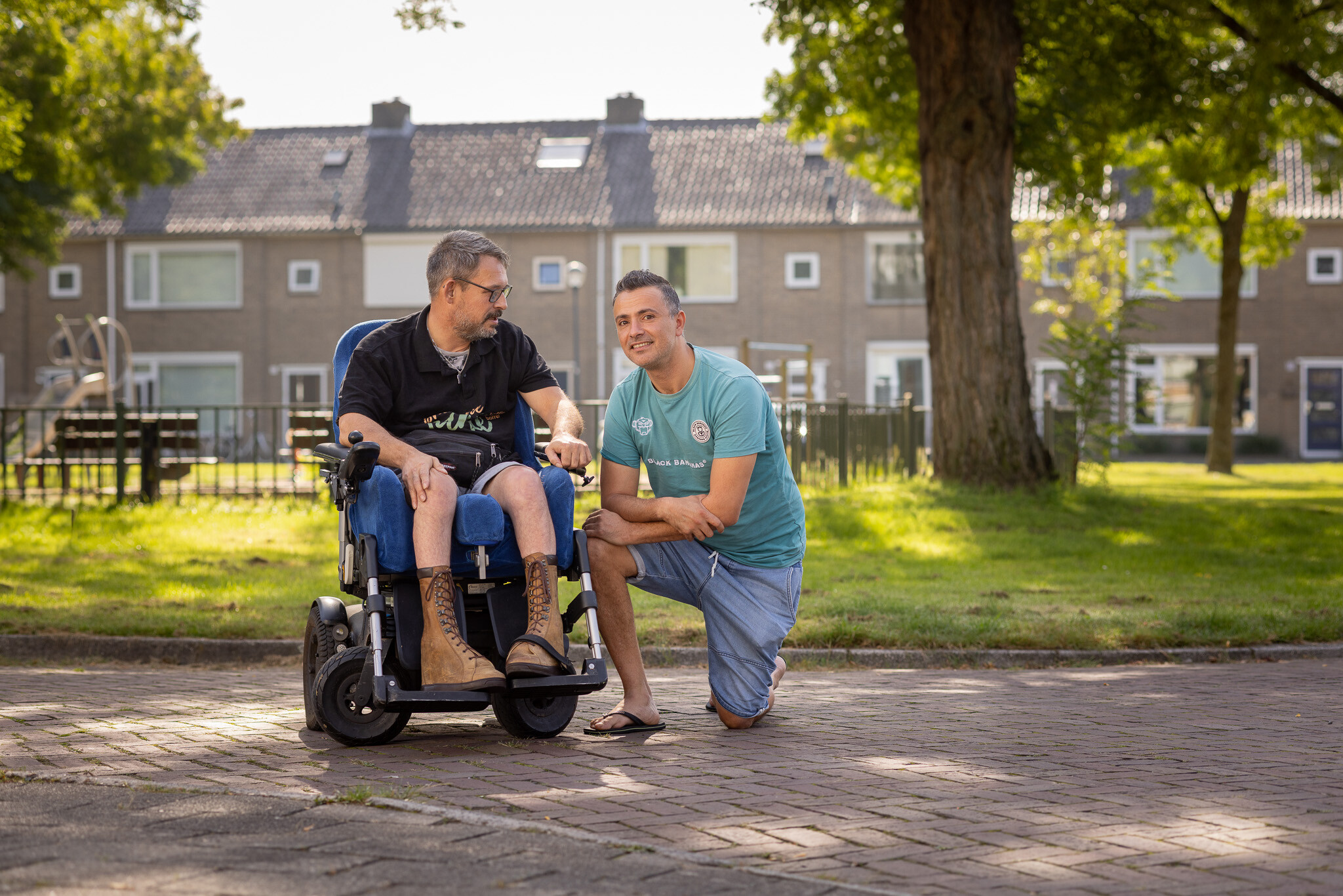 Begeleider Rachid glimlacht en knielt neer naast cliënt in rolstoel.