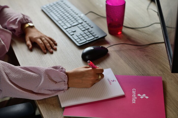 Iemand zit aan een bureau met computer en schrijft iets op in een Careflex notitieboekje